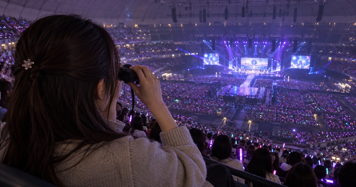 ドームのスタンド席からコンパクト双眼鏡で遠いライブステージを見る観客